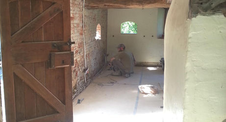 A worker crouches on a sunlit floor in a rustic hallway with exposed wooden beams and brick walls