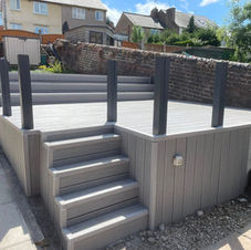 A backyard with a paved stone patio, gravel borders, raised garden beds, wheelbarrows, and a small playhouse, enclosed by a tall wooden fence with ongoing landscaping work