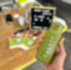 Hand holding green juice bottle labeled "Cranky Carrot." Background has a burlap tablecloth, flyers, and sign: "6 juices for 55, Kombucha cup."