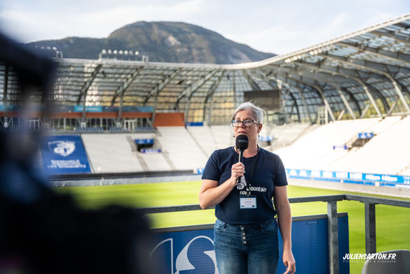 Femme avec un micro parle devant un stade de foot avec une montagne.
