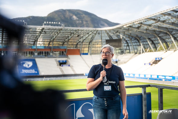 Femme avec un micro parle devant un stade de foot avec une montagne.