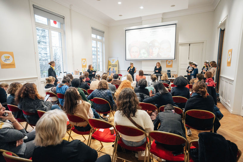 Conférence dans une salle, public assis, femmes présentes, écran projetant des images.