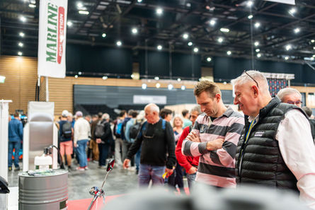 Plusieurs hommes regardent un objet, texte MARLINE. Exposition, foule, intérieur d'un hall.
