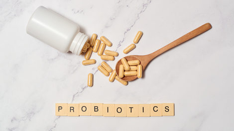 Bottle of probiotic capsules tipped onto a marble surface beside a wooden spoon filled with capsules, with the word “probiotics” spelt out in letter tiles.