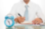 person writing at their desk with a clock in the background, symbolizing the importance of creating a routine and setting aside specific times for writing or editing.
