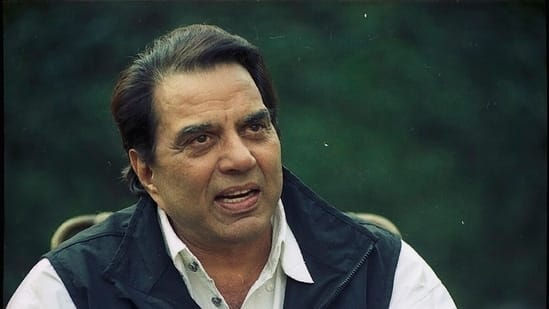 Dharmendra in a white shirt and dark vest speaking outdoors, with a blurred green background. He appears to be engaged in conversation.