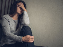 A distressed woman in a gray sweater sits against a wall, covering her face with one hand in a somber indoor scene.