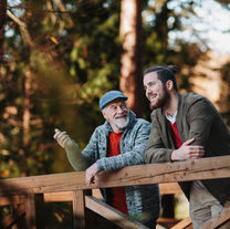 Two men, one older and one younger, smile while leaning on a wooden bridge in a forest. Both wear red shirts, creating a warm, friendly mood.