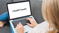 Person typing on a laptop with "ChatGPT Health" on screen, sitting on a gray couch. Casual setting, focus on hands and screen.