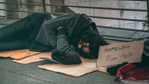 Homeless person sleeps curled on cardboard by a railing, beside a sign reading Homeless Please Help in a bleak urban scene