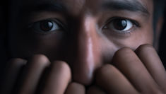 Close-up of a person's wide eyes peeking over clasped fingers in dim light, creating a tense, watchful mood.