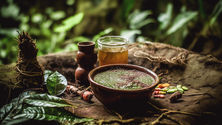 A rustic setup with a bowl of green paste, a wooden cup, a jar of liquid, and green leaves on a forest floor, creating an earthy ambiance.