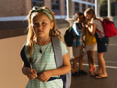 A young girl in a striped shirt stands alone, looking sad. In the background, three children whisper behind her back.