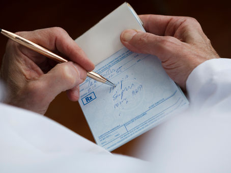 A person in a white coat writes on a blue prescription pad with a gold pen. The pad displays text and an RX symbol.