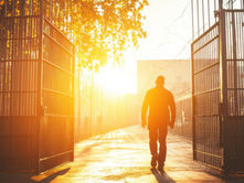 A person walks through open gates into the sunset along a fenced path.