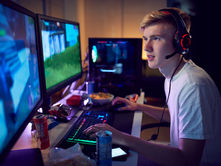 A teen wearing a headset plays video games at a dual-monitor desk, lit by neon colors, with snacks and drinks nearby.