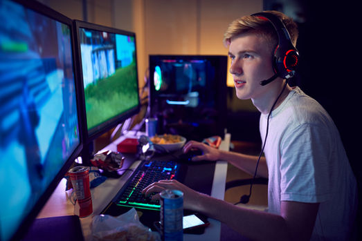 A teen wearing a headset plays video games at a dual-monitor desk, lit by neon colors, with snacks and drinks nearby.