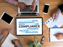 Overhead view of hands working at a table with a tablet displaying words like COMPLIANCE, charts, and notebooks. 