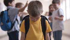 Sad boy in yellow shirt with backpack looking down, stands in school hallway as four blurred kids in background point and laugh.