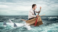 Man in a cardboard box paddles in the ocean, surrounded by shark fins.