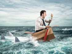 Man in a cardboard box paddles in the ocean, surrounded by shark fins. 