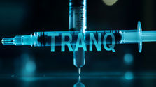 A syringe with blue liquid, needle tip down, glows against a dark background with the word "TRANQ" overlaid, a nickname for the drug Xylazine.