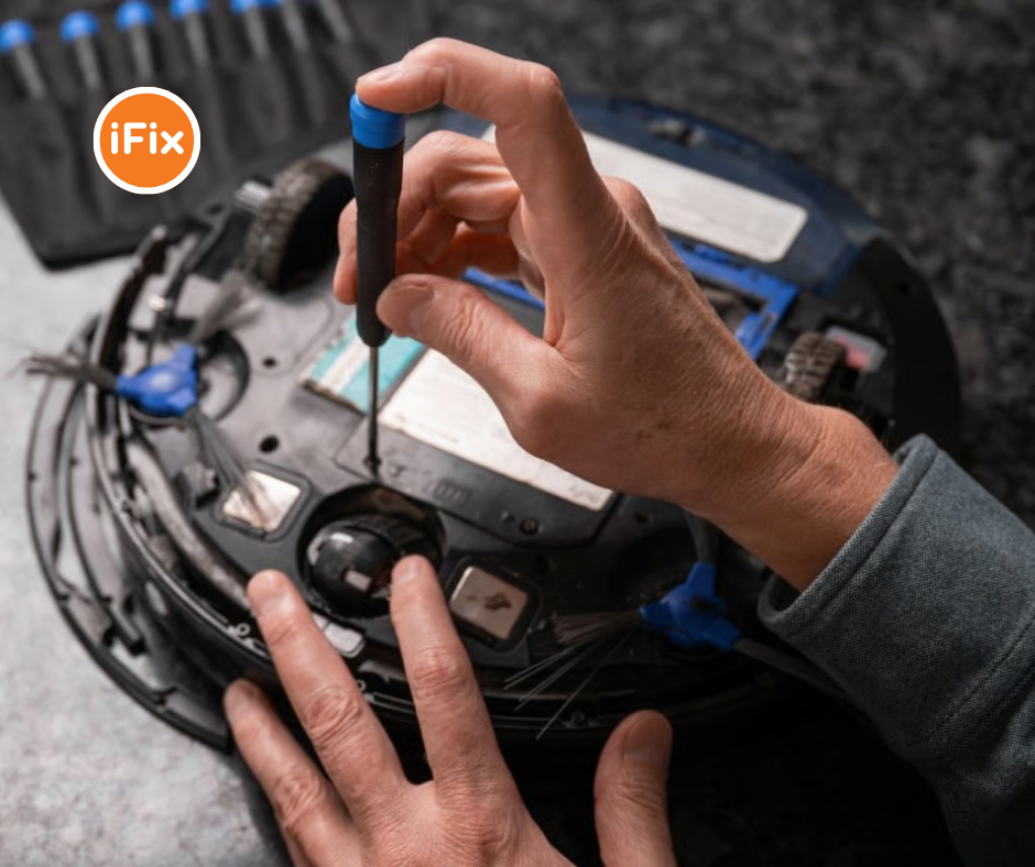 A person using a precision screwdriver to repair the underside of a robot vacuum cleaner, with tools visible in the background and the iFix logo in the upper left corner.