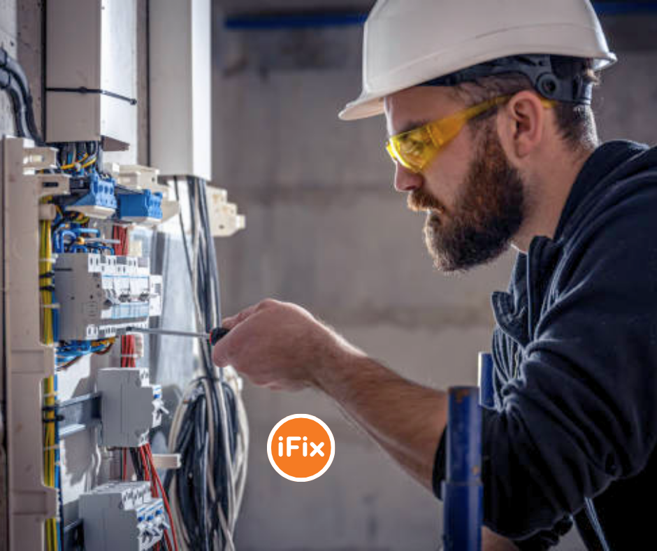 A bearded electrician wearing a white hard hat and yellow safety glasses carefully working on a wiring panel, with the iFix logo overlaid in the foreground.