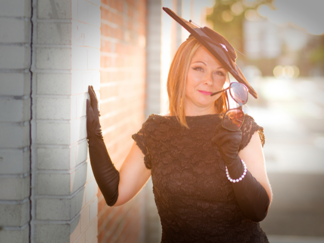 Woman wearing a 1940s style dress and hat, leaning on a wall in the sun