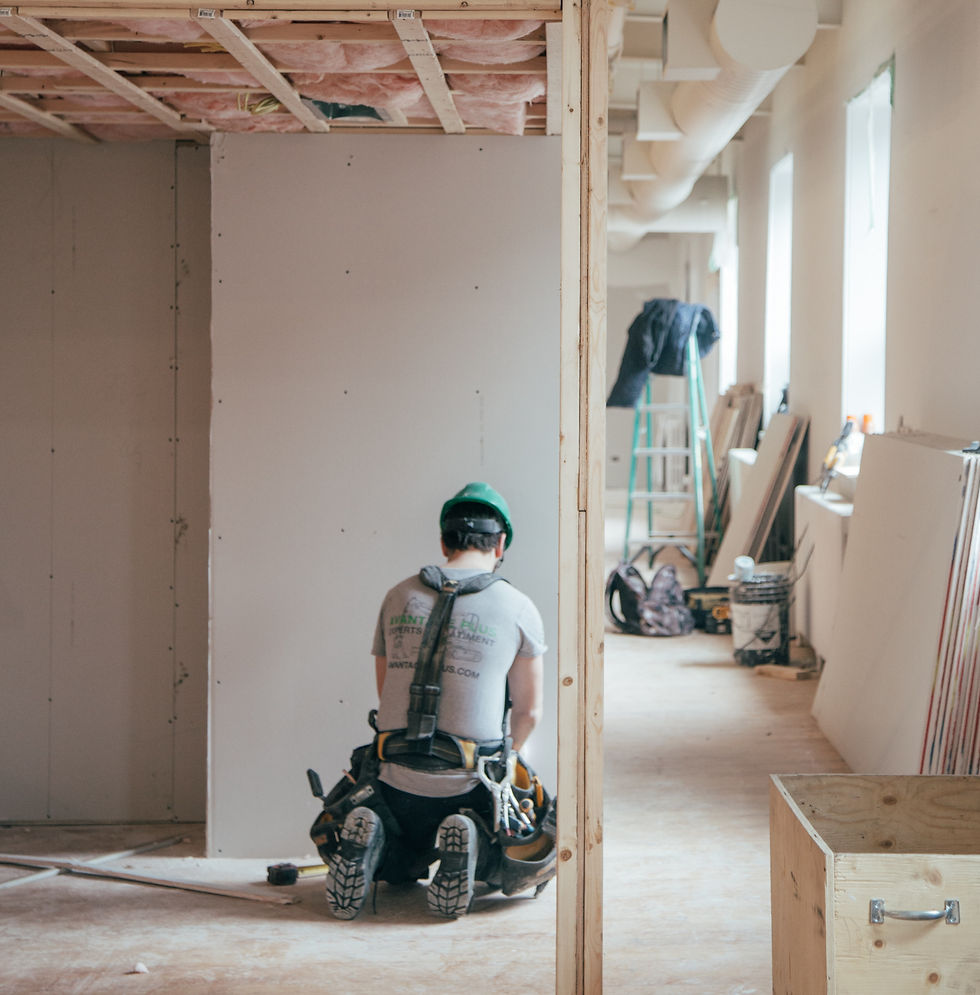Worker kneels installing drywall in a room under construction. He's wearing a green helmet. Tools and materials are scattered around.