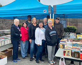 Group of 5 women and men standing with books on tables under a blue canopy