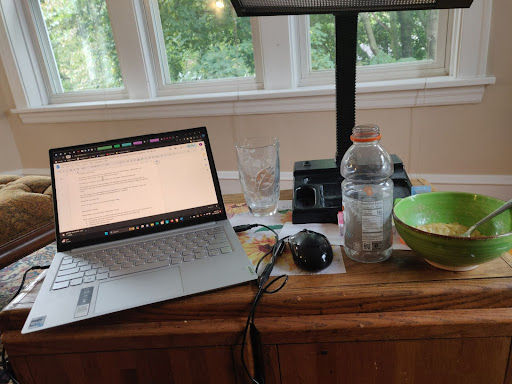 Photo showing a low cabinet in front of a window that’s being used as a makeshift table. A laptop takes up most of the space on the placemat, along with an empty glass. A lightbox, pill bottle, and empty Gatorade bottle fill most of the remaining space. A bowl of food is smushed into the corner.