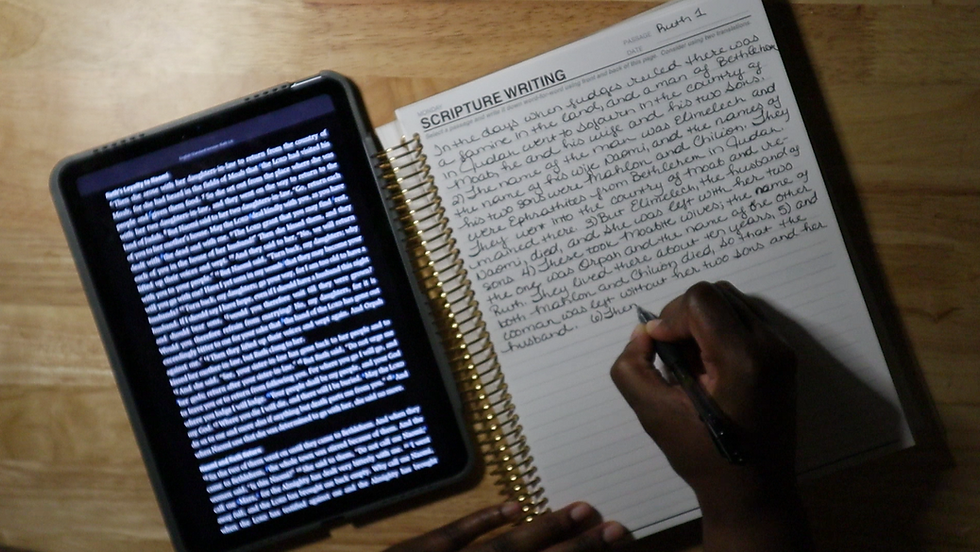 A person's hand holding a pen writes in a spiral-bound Scripture Writing journal, copying the book of Ruth chapter 1 in neat cursive handwriting. A tablet displaying the biblical text in white text on a dark background is propped open beside the notebook on a wooden surface, serving as the reference for transcription.