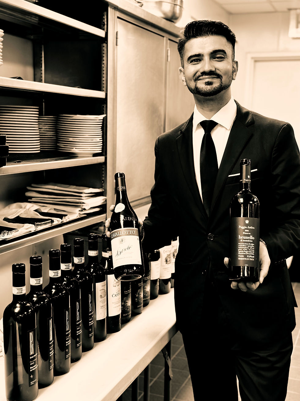Man in a suit holding two wine bottles, standing in a room with shelves of plates and bottles. Black and white image with a confident mood.