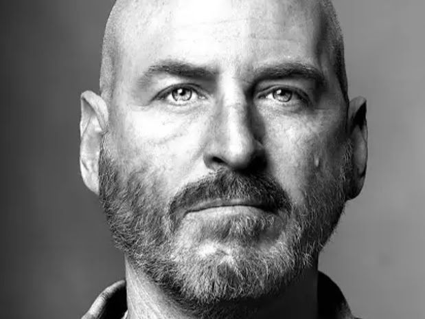 Britton Day with beard in grayscale portrait. Serious expression, neutral gray background, focused gaze.
