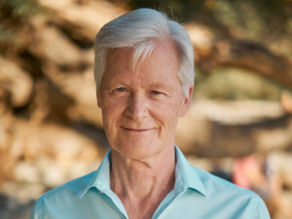 Udo Erasmus with white hair smiles softly, wearing a light blue shirt. Blurred natural background of trees suggests an outdoor setting.