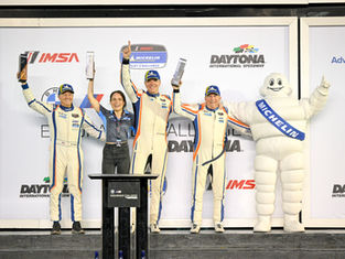Ted Giovanis and Team TGM celebrate a class win on the Daytona podium alongside co-drivers and the Michelin Man mascot, all holding trophies and raising their arms in victory after the 2026 season opener.