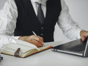 Executive writing notes in a notebook beside a laptop while planning or reviewing priorities at a desk.