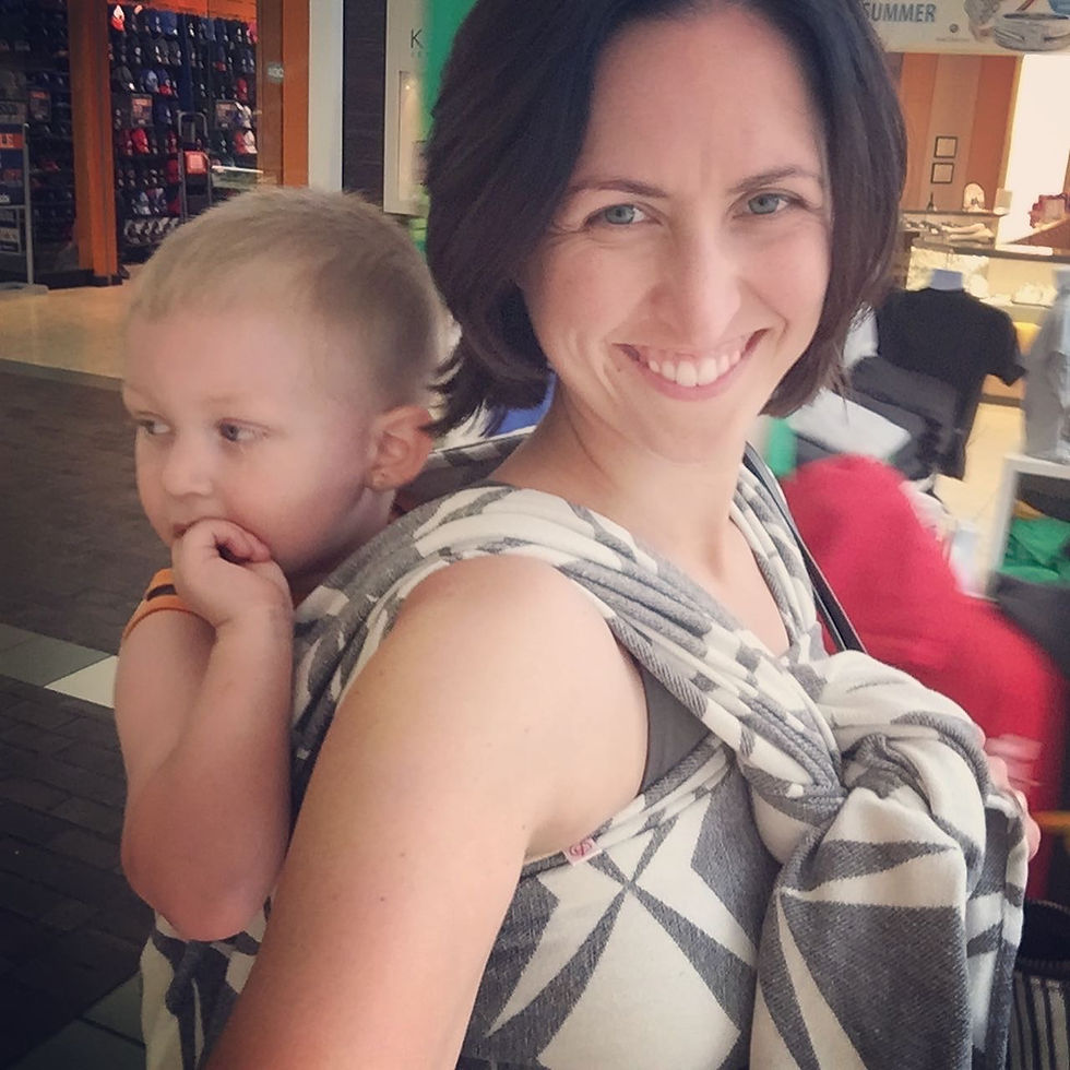 Woman smiling with a child on her back in a patterned sling at a shopping mall. Bright setting, child looks thoughtful.