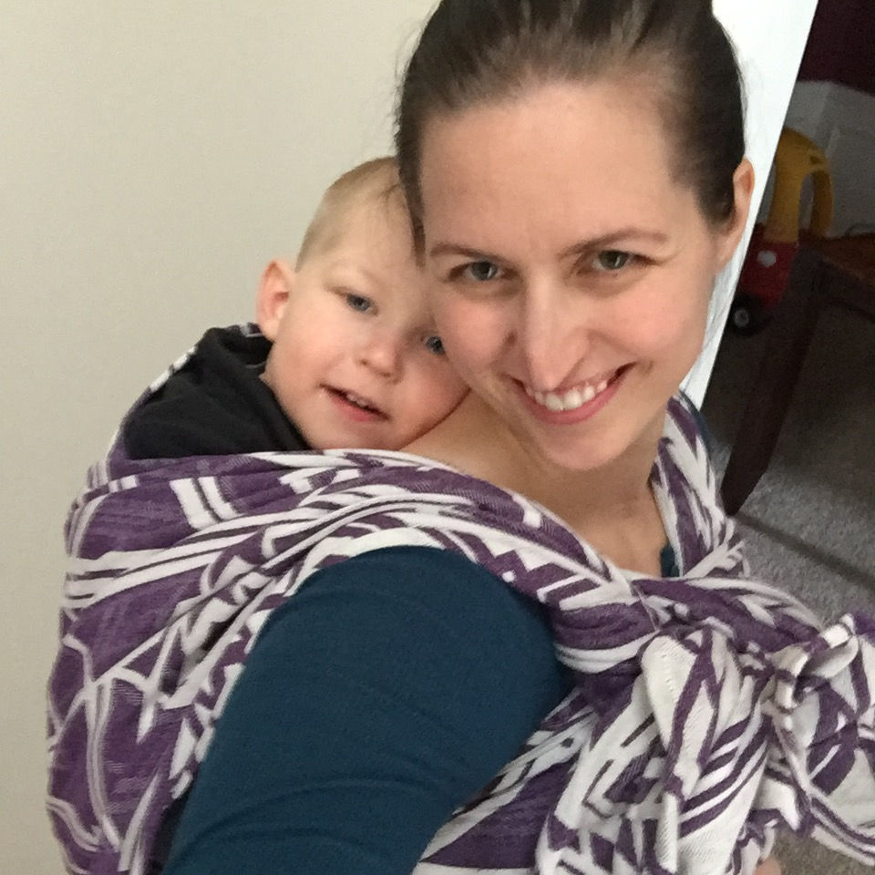 Smiling woman carries a baby in a purple and white wrap. Both look content. Indoor setting with a toy and neutral walls.