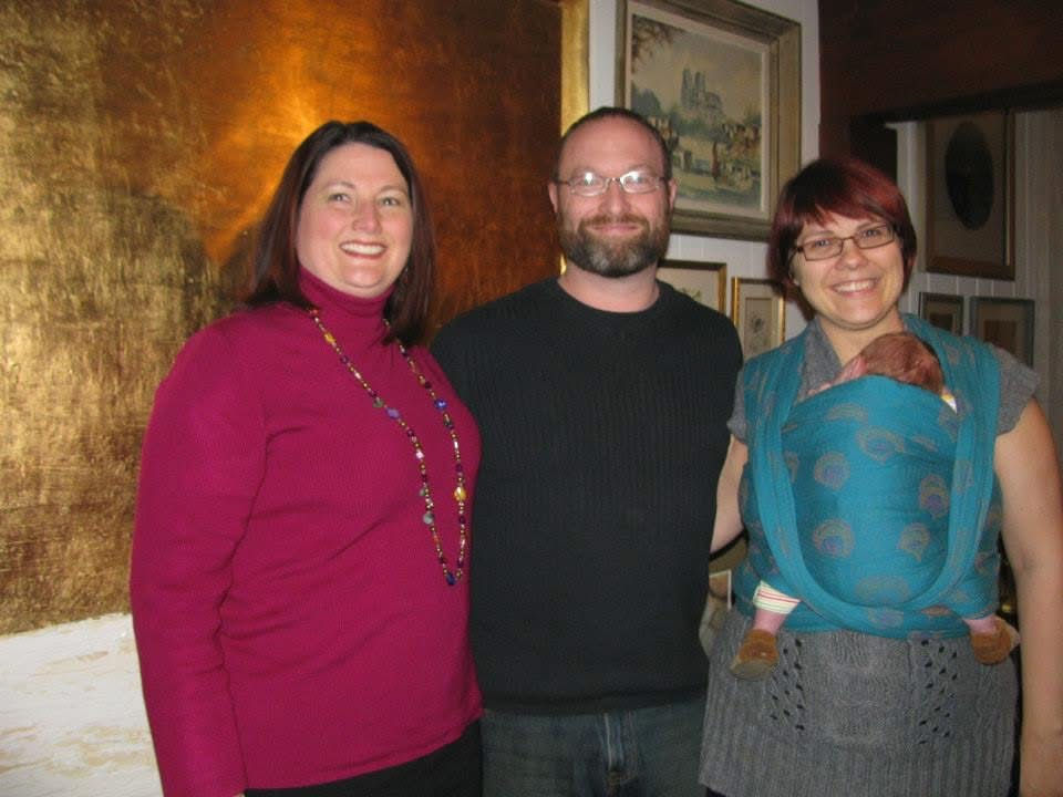 Three adults smiling indoors, one woman with a baby in a blue baby carrier wrap. Warm lighting, gold wall, and framed pictures in the background.