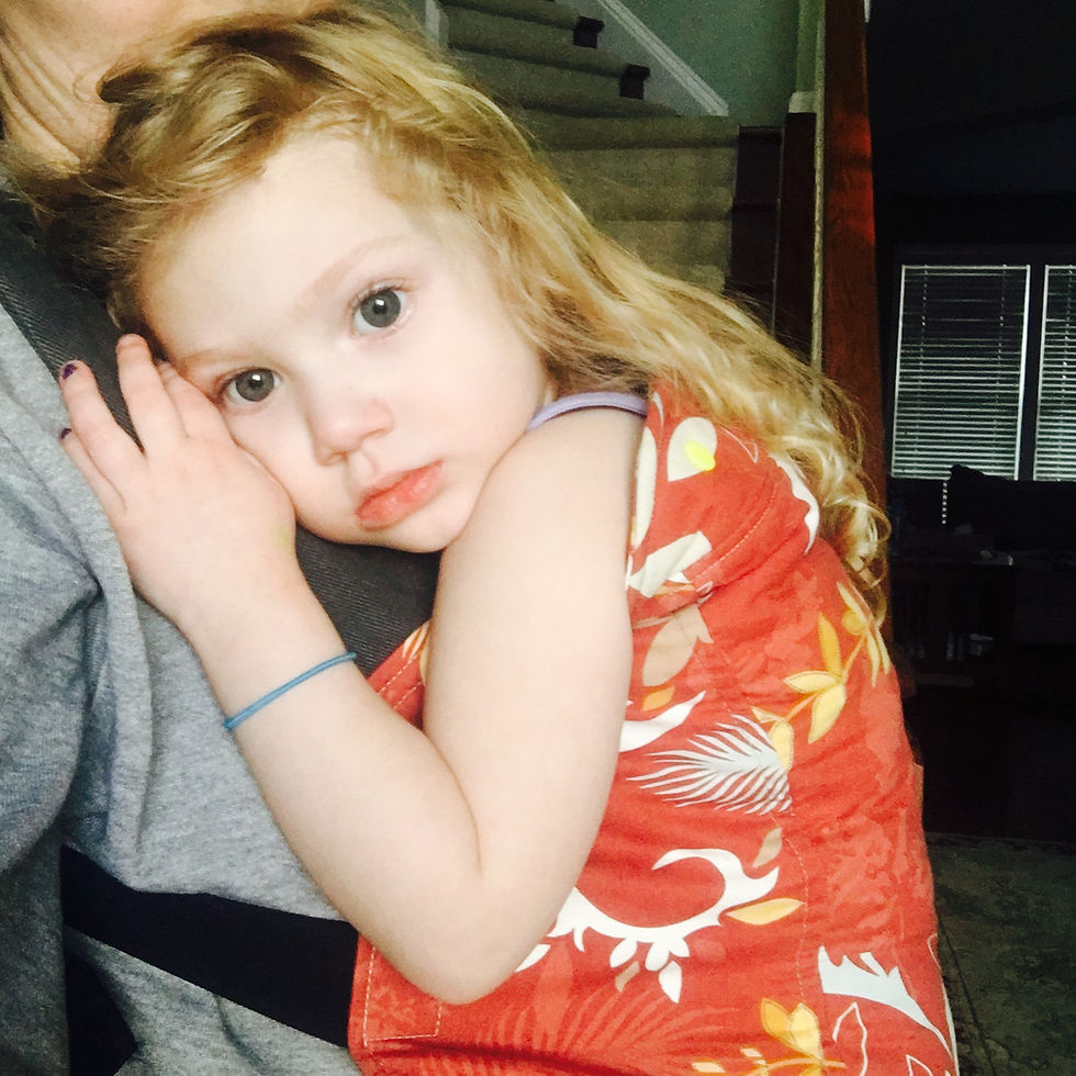 Child with long blond hair in a red floral dress rests against an adult in a baby carrier. Calm expression, indoor setting with stairs and dim background.