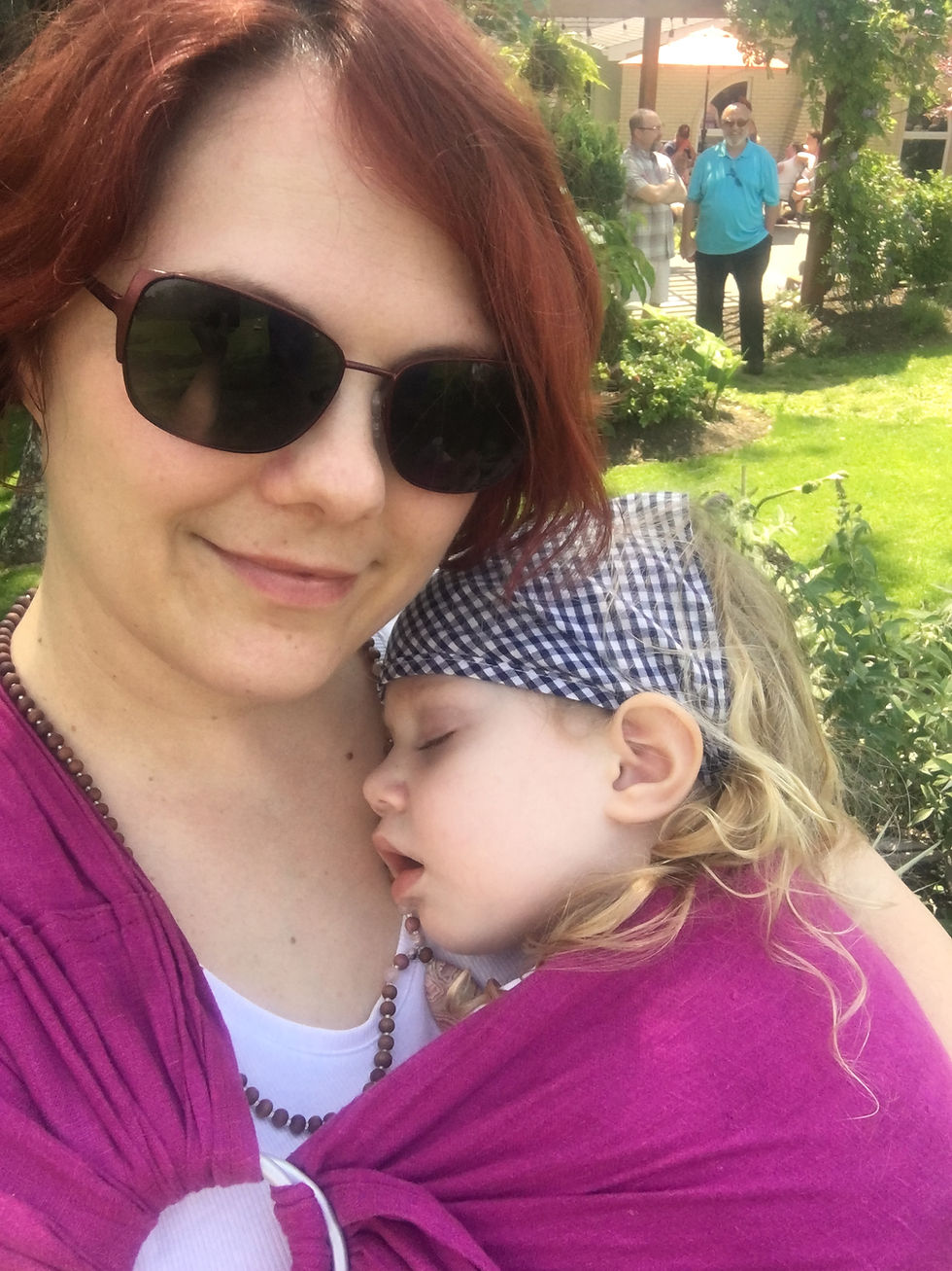 Woman in sunglasses holds sleeping baby in pink wrap outdoors. Greenery and people in background. Peaceful mood.