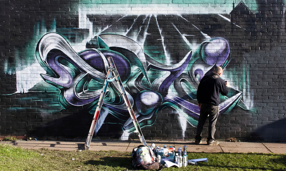 A man painting graffiti onto a wall that is coloured green, purple and silver