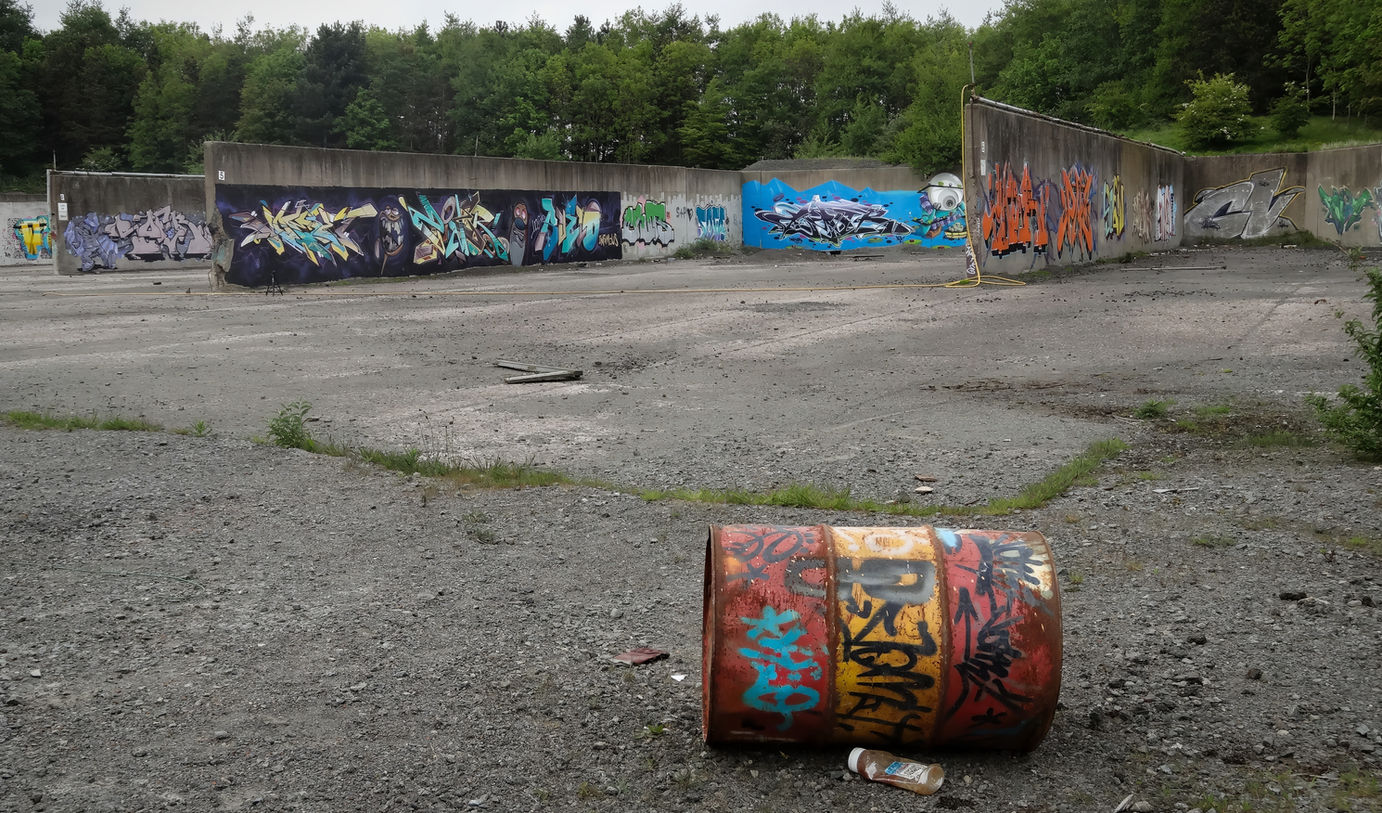 An old oil can or barrel, covered in graffiti tags, with walls full of graffiti behind it