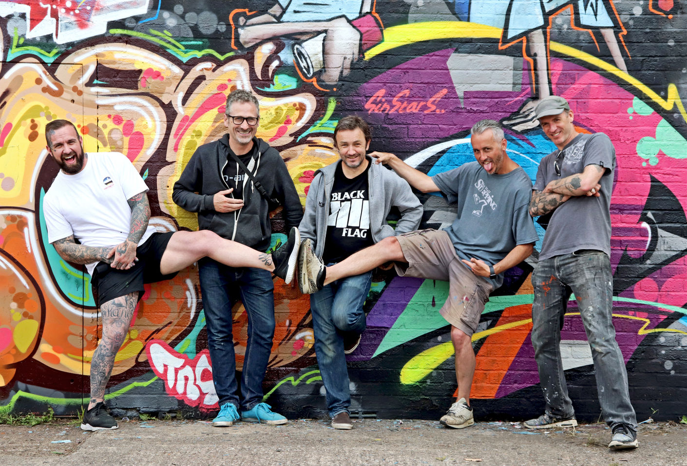 Five graffiti artists posing in front of a wall of graffiti that they'd just spray painted