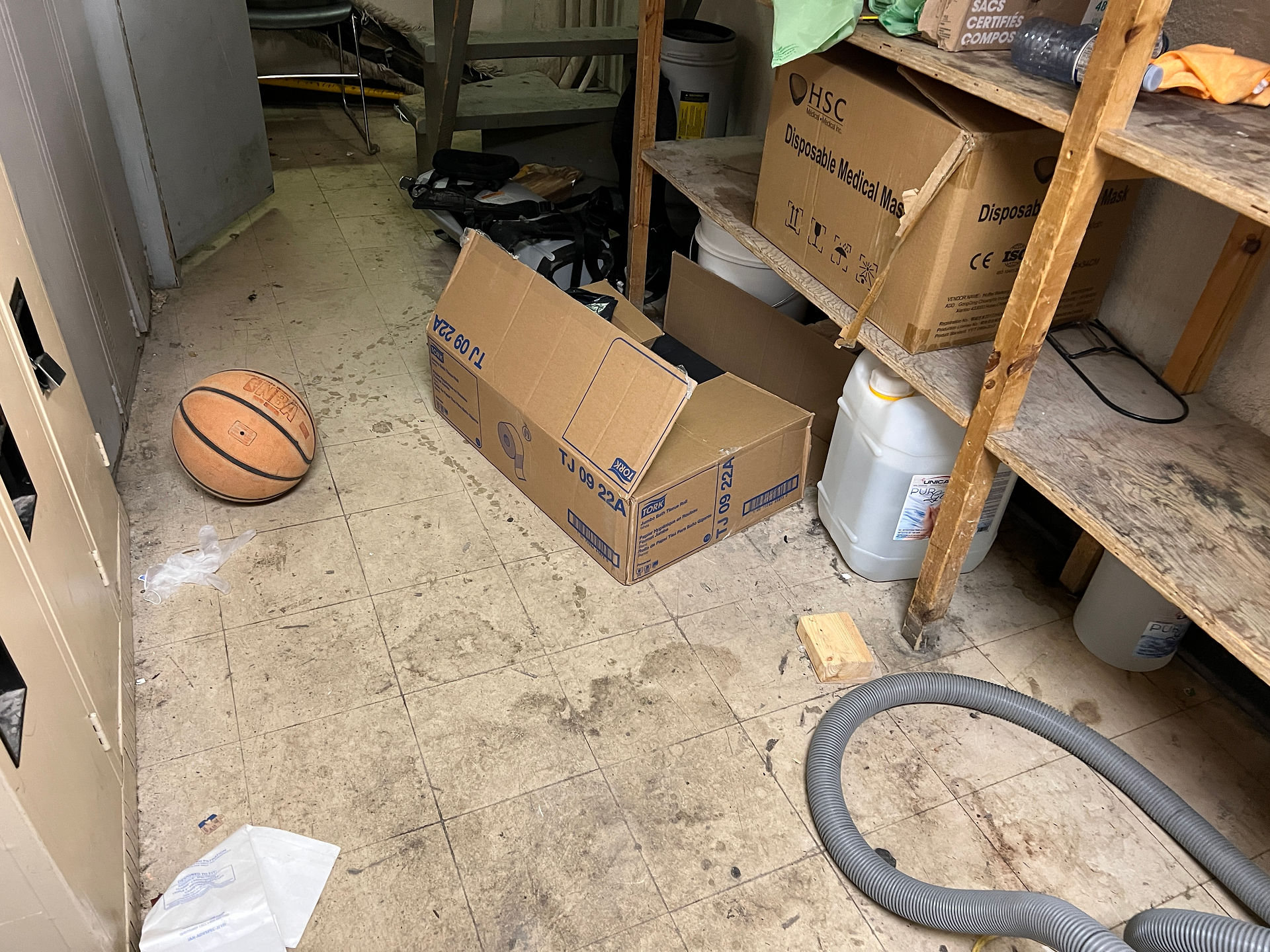 Local de conciergerie encombré avec sol sale, boîtes de carton au sol, équipement mal rangé, présence d’objets personnels comme un ballon de basketball, illustrant un manque de professionnalisme et de zonage clair.