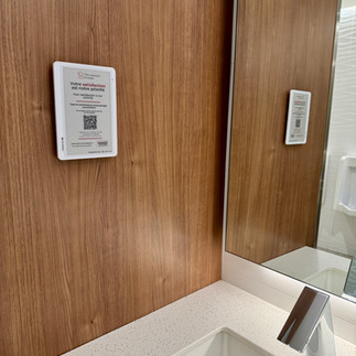 Borne Service+ installée sur un mur en bois dans une salle de bain, affichant un message de satisfaction et un QR code, avec son reflet visible dans le miroir au-dessus du lavabo.