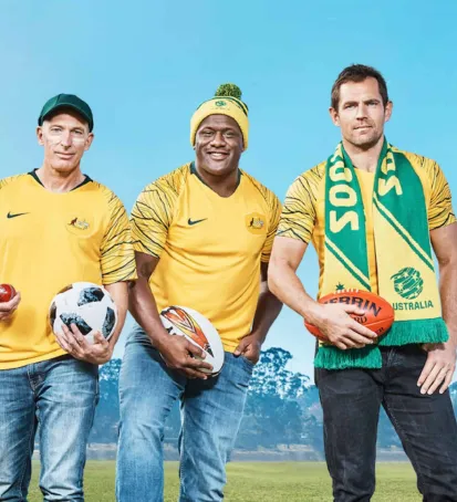 The image features three athletes representing different sports, standing together in a sports field setting. They are all wearing yellow sports jerseys with the logo of Australia on the chest.

From left to right:

The first person is holding a cricket ball and a soccer ball, representing cricket and soccer.
The middle person is wearing a beanie, holding a rugby ball, symbolizing rugby. He has a broad smile, adding a friendly vibe to the group.
The third person is wearing a green and yellow scarf with "Socceroos" written on it and is holding an Australian rules football, indicating support for the Australian football team.
The background is clear with a blue sky, suggesting an outdoor setting. The athletes' outfits and props emphasize national pride and sports diversity in Australia.