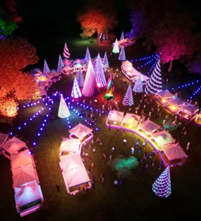 A vibrant aerial image of an outdoor nighttime event featuring illuminated tents, cone-shaped light installations, and colorful lights decorating the area. People are gathered around the glowing tents, which form a winding path through the venue. The trees surrounding the event are lit up in various colors, adding to the festive atmosphere. The illuminated cones display different patterns and colors, creating a magical and whimsical environment that suggests a festival or celebration.






